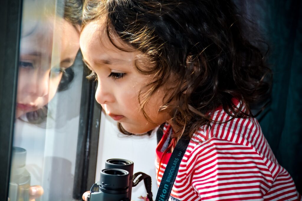 Young girl with a stripped shirt holding binoculars and looking out a window being curious about the world around her.